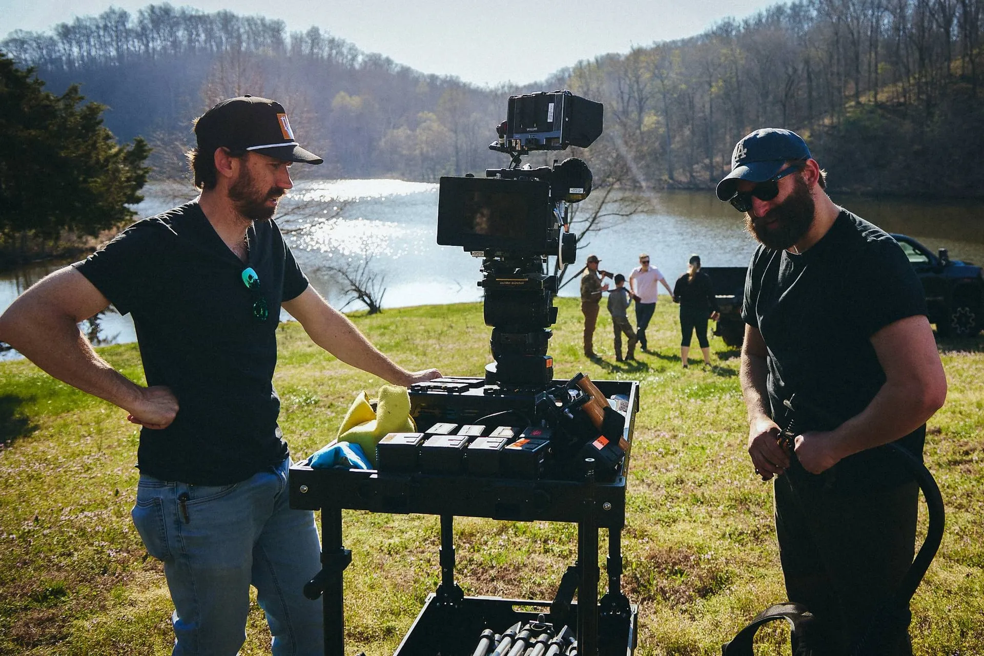 Two crew members beside a cinema camera on a cart, a lake and wooded hills behind them, a family standing at the water’s edge.