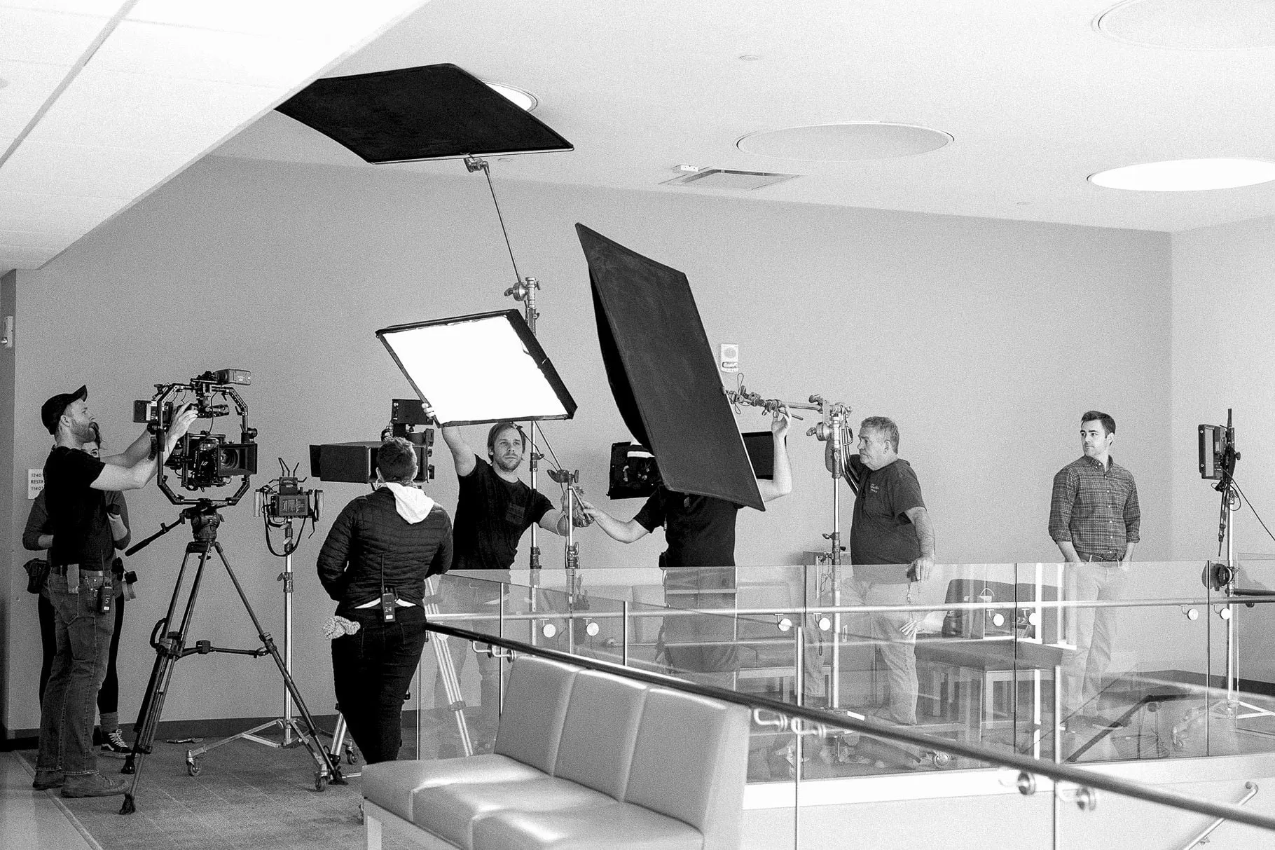 A crew in black and white lifting flags and lights into place on a glass-railed mezzanine while the camera stands ready.