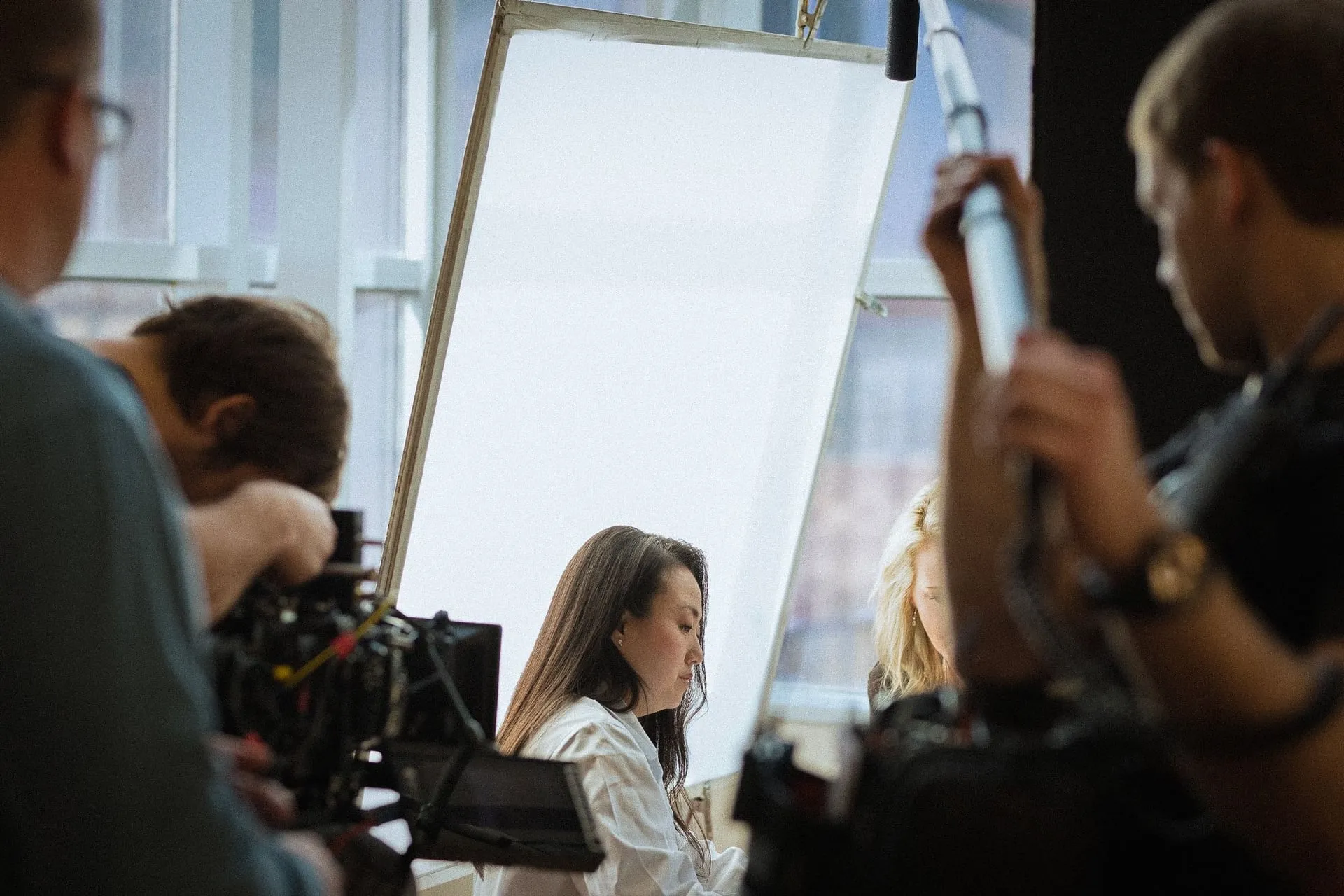 A woman in a white coat looks down, framed between two camera operators and a large diffusion frame.