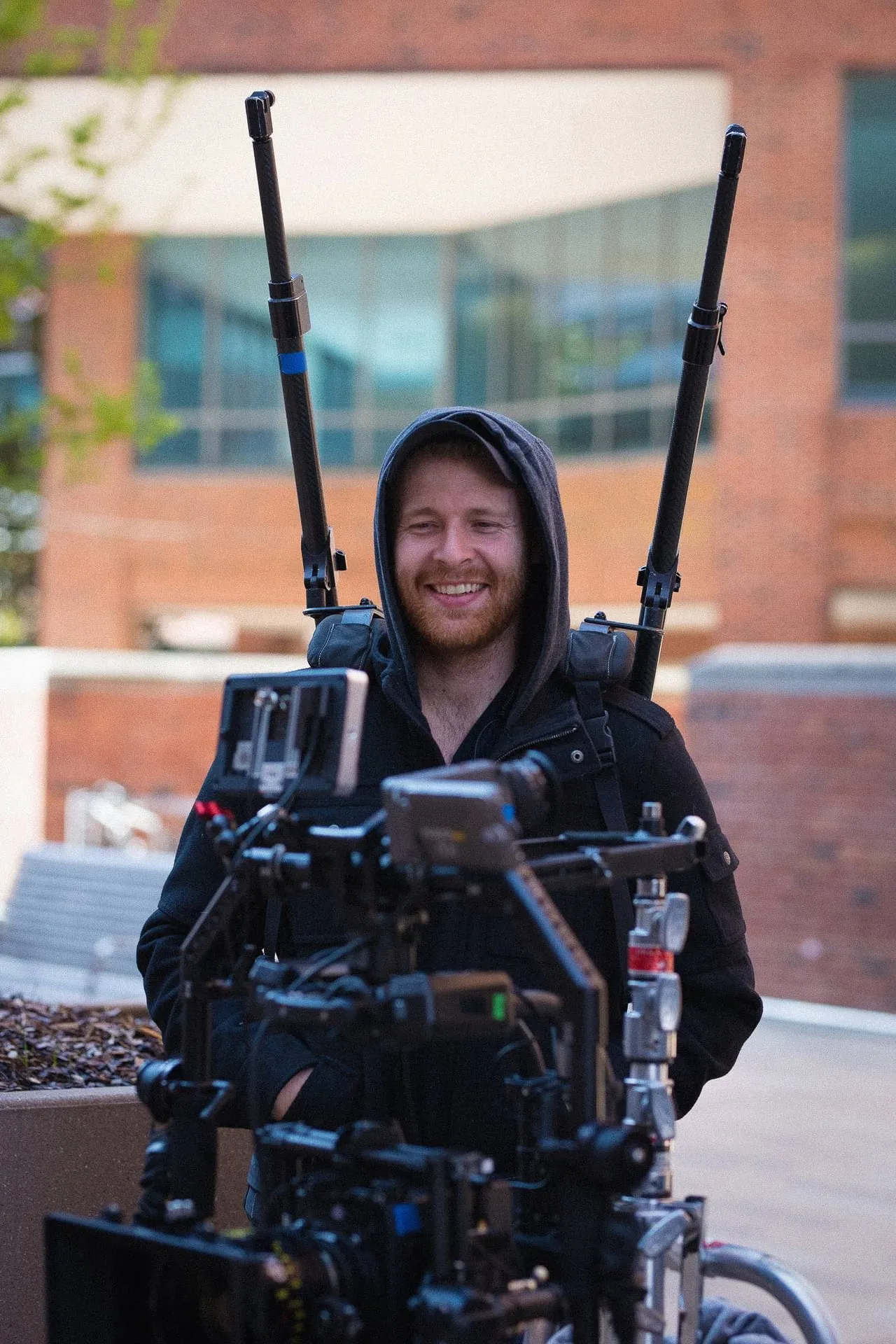 A smiling crew member in a hooded jacket stands behind a camera rig with two boom poles on his back.