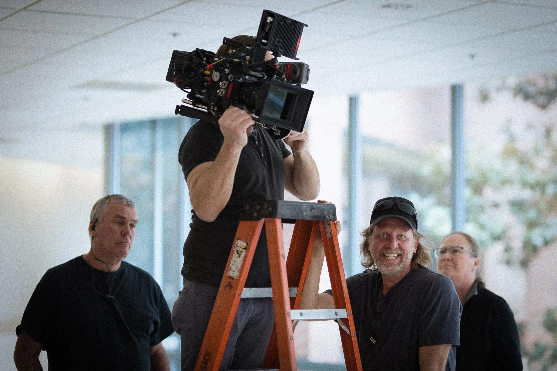 A camera operator on a stepladder with the camera on his shoulder, two crew members watching and one grinning up at the lens.
