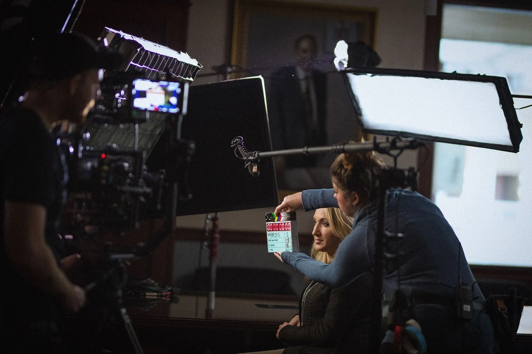 A crew member holds up the slate in front of a seated interview subject, with a camera, light and boom microphone all around.