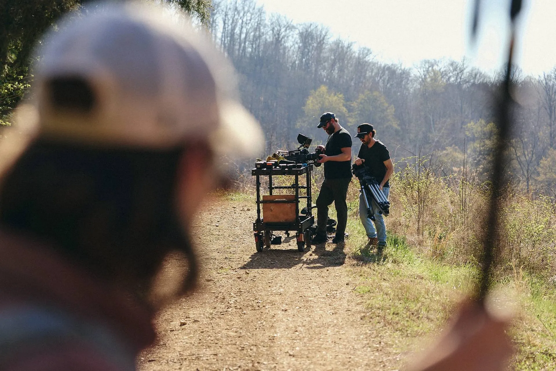 Two of our crew ready a cinema camera and tripod at the camera cart on a dirt trail, a third crew member out of focus in the foreground.