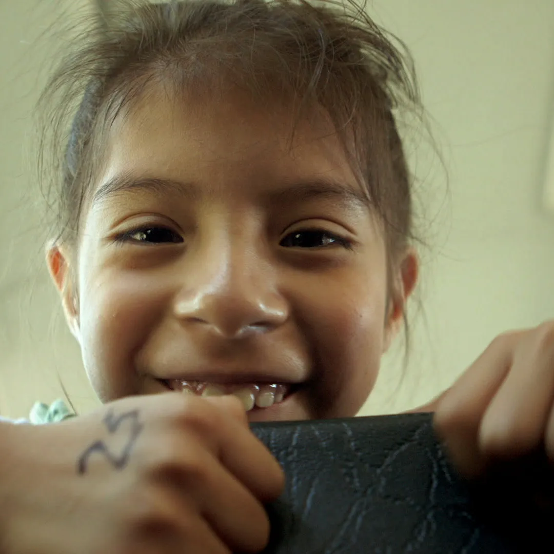 A young girl grins over the back of a bus seat, a number written on her hand.