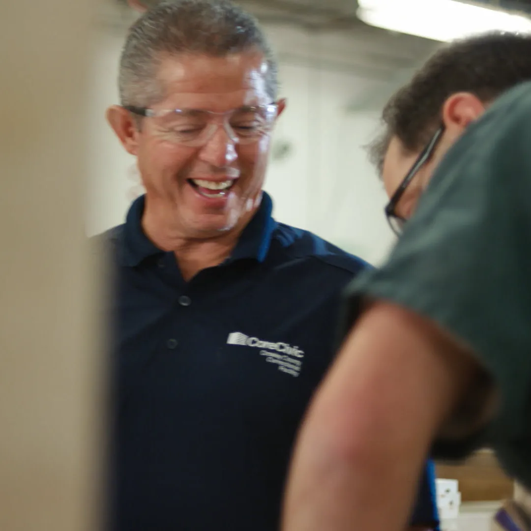 A CoreCivic instructor in a navy polo laughs as he works beside a man in a green uniform.