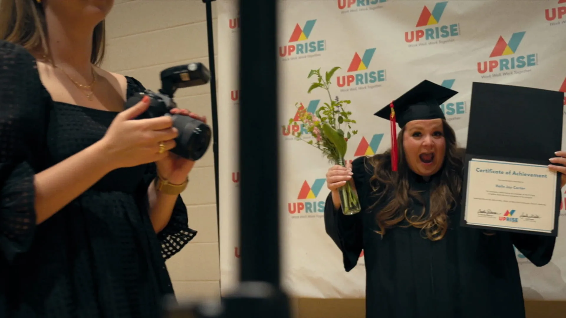 A graduate in cap and gown beams as she holds up her certificate for a photographer.