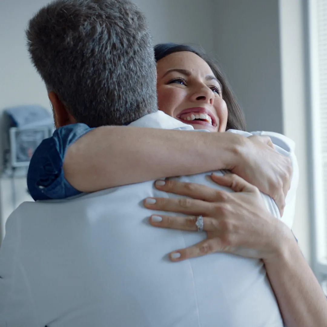 A woman laughs as she hugs a doctor in a white coat in an exam room.