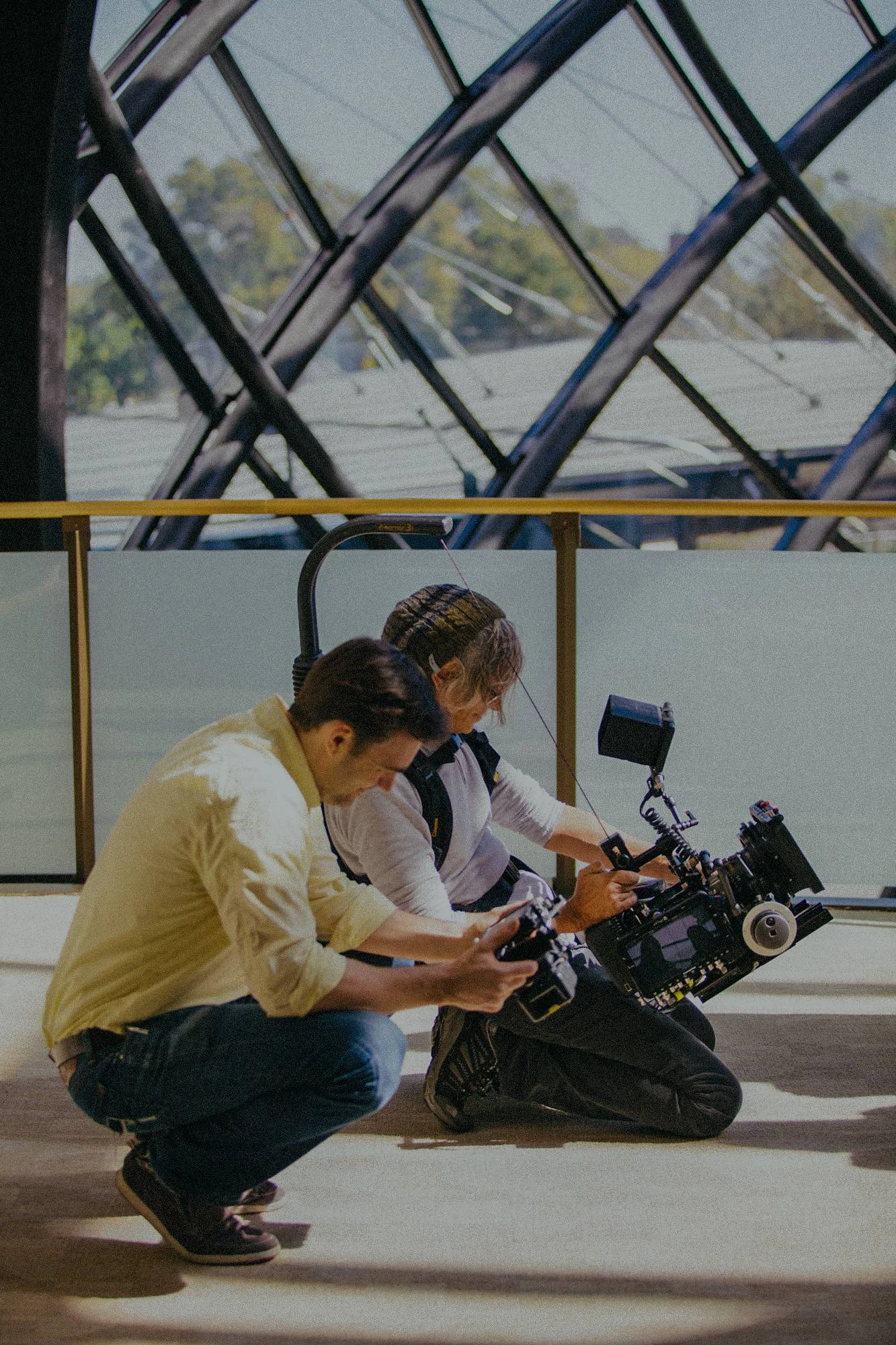 Two crew members crouched on a carpeted floor in bright window light, checking a cinema camera rig, with a large diamond-lattice window behind them.